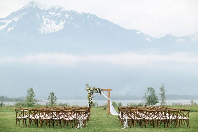 An empty outdoor ceremony setup with wooden chairs, a floral arch, and a misty mountain backdrop